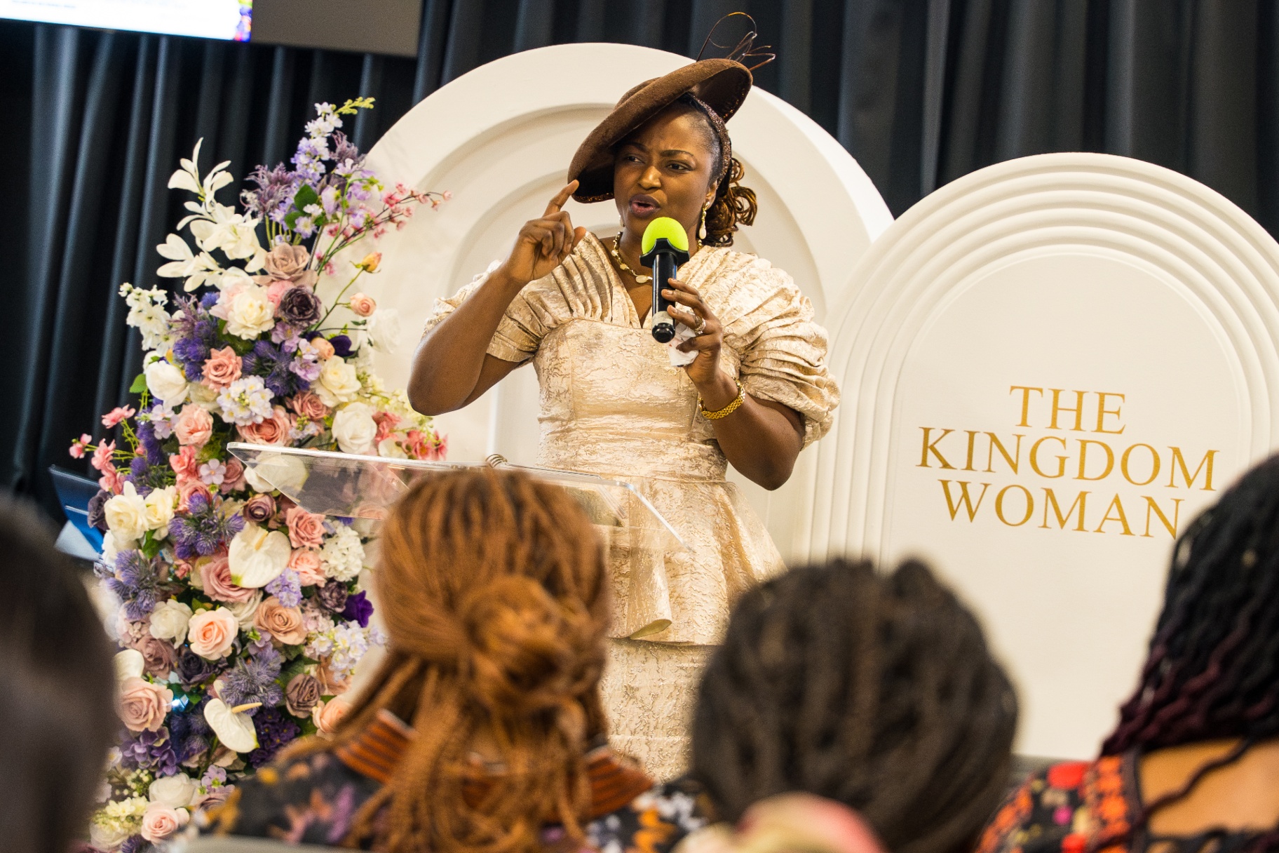 Speaker in cream dress and hat preaching at The Kingdom Woman