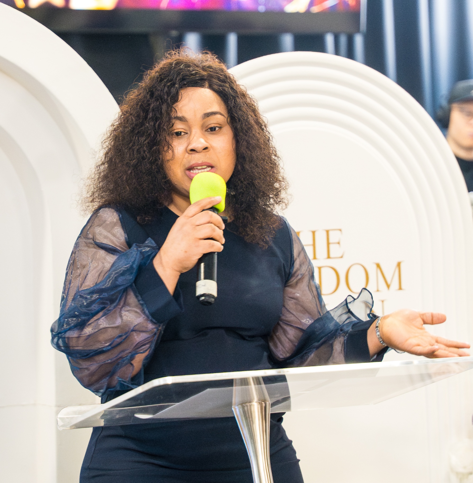 Speaker in navy sheer-sleeve dress ministering at The Kingdom Woman